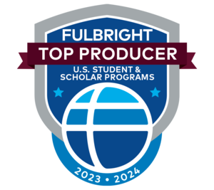A badge consisting of a bright blue shield overlaid with a dark red ribbon near the center and a stylized globe at the bottom. Text reads: Fulbright Top Producer / U.S. Student Program / 2023-2024