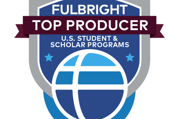 A badge consisting of a bright blue shield overlaid with a dark red ribbon near the center and a stylized globe at the bottom. Text reads: Fulbright Top Producer / U.S. Student Program / 2023-2024