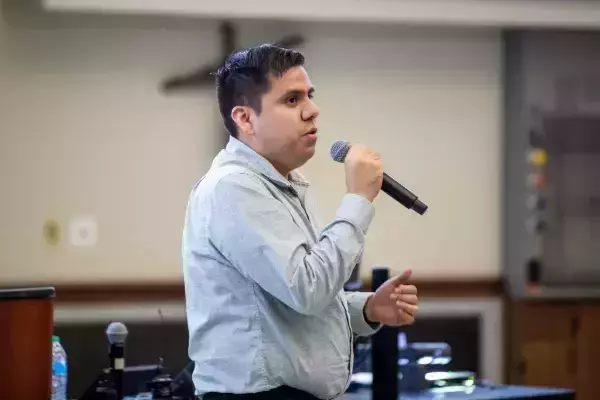 A man with dark hair wears a light blue shirt, speaking intently into a black microphone. His left hand is slightly raised in a gesture, as he stands in a lecture hall.