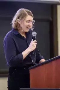 A female presenting person in a blue business suit gestures while addressing a classroom.  She stands behind a wooden podium with two computer monitors.  The audience is slightly out of focus in the foreground.