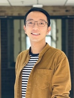 Young Asian man with glasses, wearing a brown shirt over a striped tee, blue jeans, and beaded bracelets, smiling in a hallway.