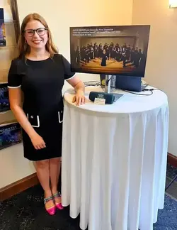 Smiling woman in black dress with white trim, glasses, by table. Monitor shows 'GAYLE RHODES and Concordia Vocal Ensemble.'