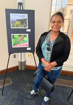 Woman in studded jacket smiles beside an easel with art: a mossy cliff landscape and a grasshopper on a pink surface.