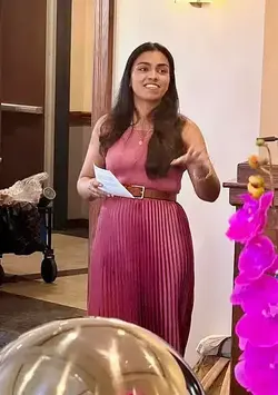 Smiling woman with long dark hair in a pink pleated dress and brown belt speaks, holding papers and gesturing.