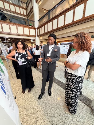A young man braided hair wearing a gray suit gestures toward a research poster as he speaks with two women at a university poster session. They stand near display boards.