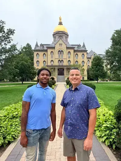 Two men smile on a brick path, the Main Building and Golden Dome of Notre Dame behind them. One wears a blue polo, the other a patterned shirt.